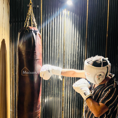 man wearing rival white color headguard and rivals boxing gloves punching the leather punching bag under spot light. bag looks high quality and is made up of leather. the impact of punch can been seen on punching bag. punching bag is held with golden chains. vintage leather heavy punching bag hung with golden chains infront of black and gold background. Cowhide leather used for heavy duty training for boxing and mma practice. High quality image of punching bag for home gyms, commercial gyms. one of a kind l