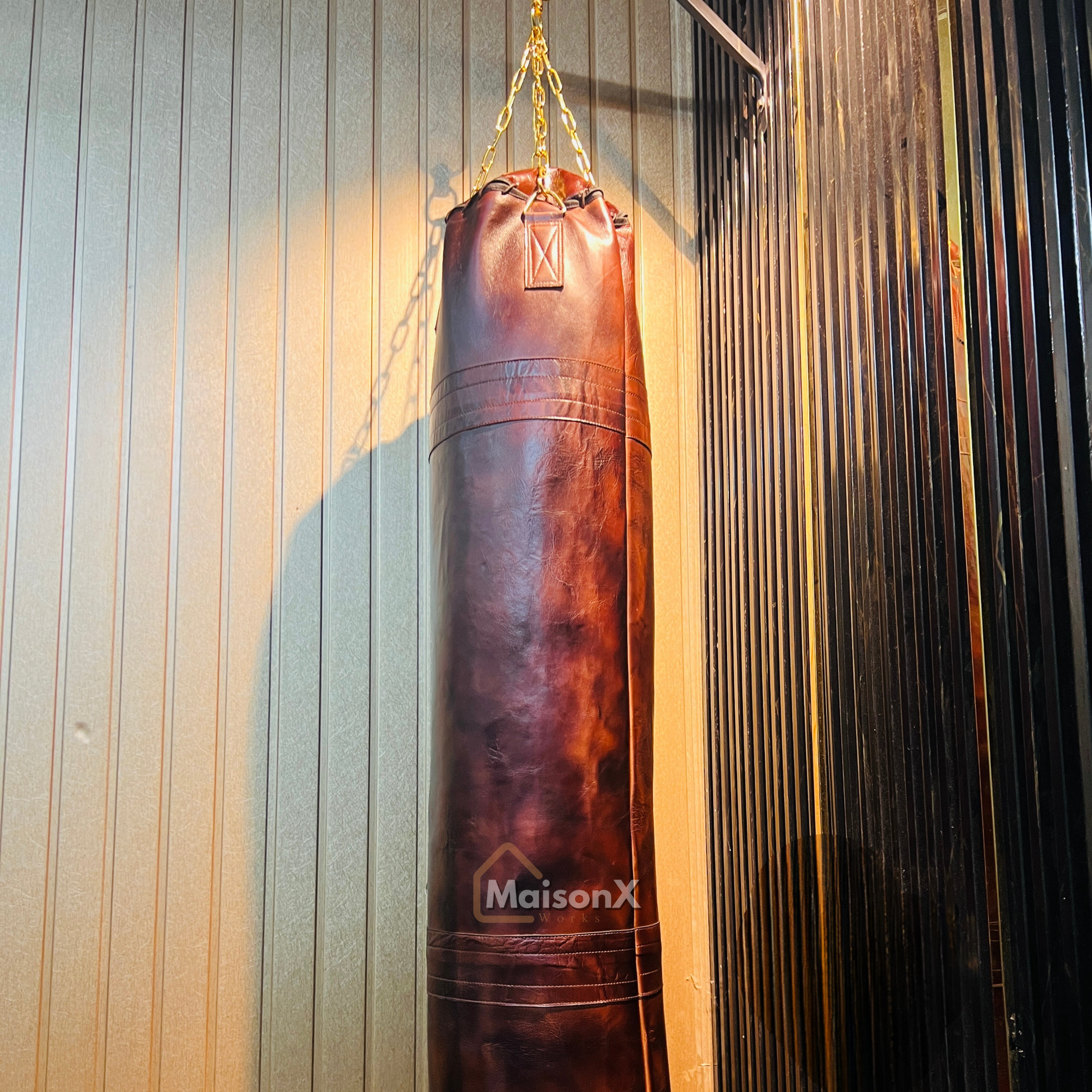 vintage leather heavy punching bag hung with golden chains infront of black and gold background. Cowhide leather used for heavy duty training for boxing and mma practice. High quality image of punching bag for home gyms, commercial gyms. one of a kind leather punching bag. brown burgundy coloe leather heavy bag for heavy duty training that stands the test f time. punching bag hanging with golden chains infront of gold and black wall