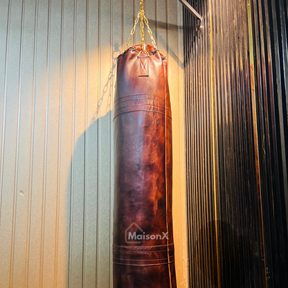 vintage leather heavy punching bag hung with golden chains infront of black and gold background. Cowhide leather used for heavy duty training for boxing and mma practice. High quality image of punching bag for home gyms, commercial gyms. one of a kind leather punching bag. brown burgundy coloe leather heavy bag for heavy duty training that stands the test f time. punching bag hanging with golden chains infront of gold and black wall
