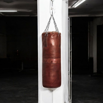 A large Blood-Fang Pro Punching Bag (up to 6 feet) hanging by its professional steel chain for a full-body MMA Kickboxing Bag or Boxing Training Equipment session. The heavy-duty cowhide genuine leather heavy bag is shipped as an unfilled punching bag, allowing customization. This handmade heavy bag is essential Gym Workout Gear and a classic vintage leather punching bag for any combat sport